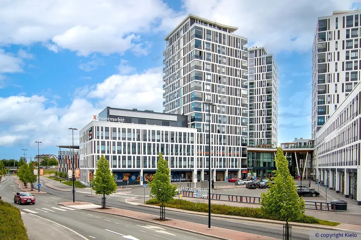 Toimitila- ja asuinkiinteistö Puutarhakatu 11, joka joka on osa Kuopion Portti / Matkakeskus Kuopio kiinteistökokonaisuuutta. Talossa valkea julkisivu ja paljon ikkunoita. Taustalla sininen taivas.