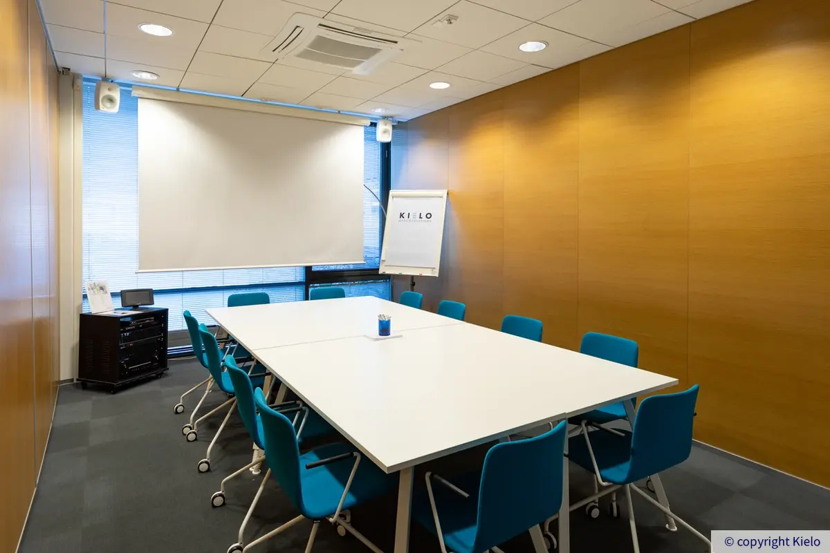 Innova 2, toimitilakiinteistön neuvotteluhuone. Kuvassa vaalea pöytä jonka ympärillä sinisiä tuoleja. Pöydän takana isot ikkunat sekä fläppitaulu