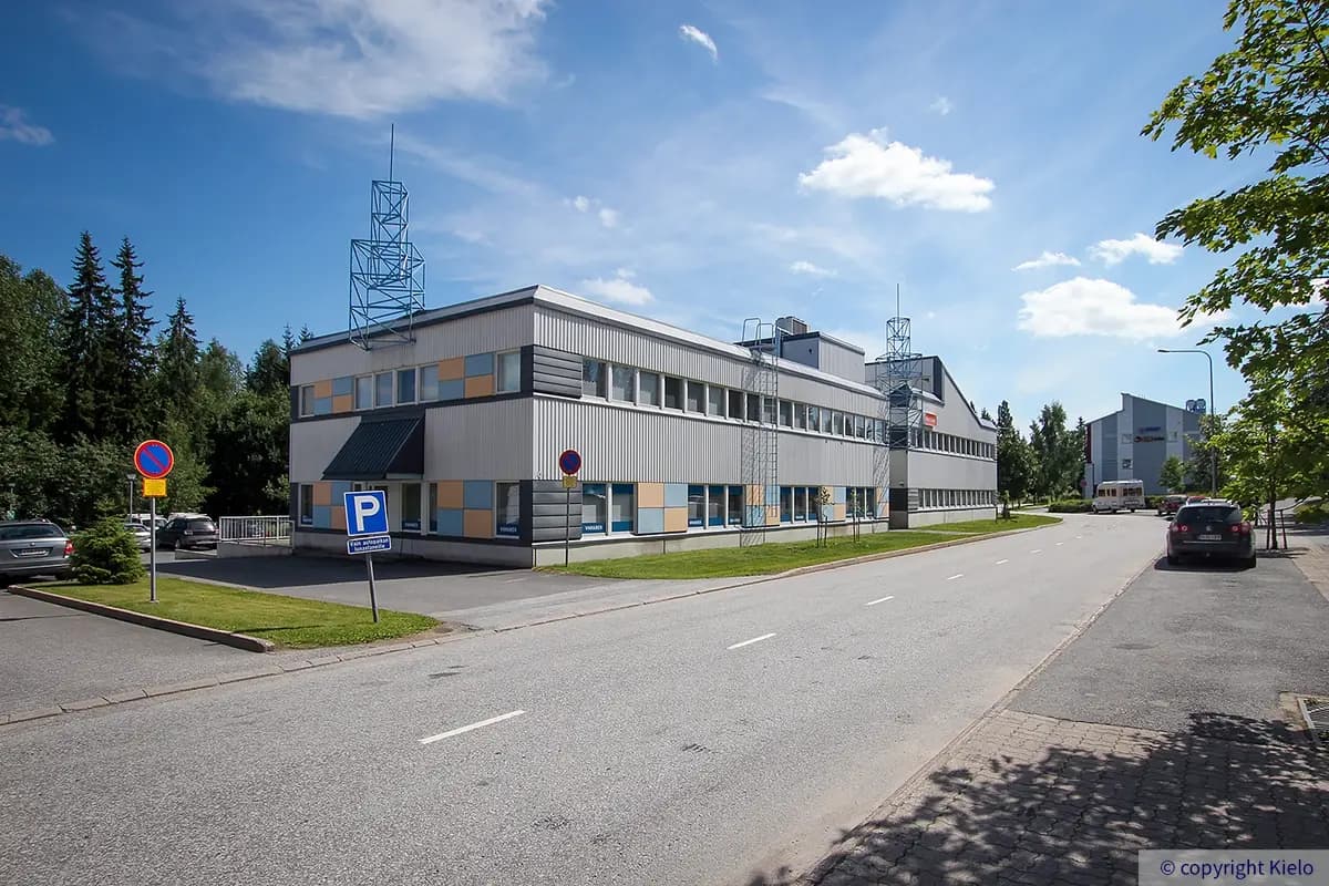 Jyväskylän Ylistönmäentie 24 kuvattuna kadun puolelta. Talon julkisivussa monivärisiä paneeleja. Taustalla metsää, taivaalla poutapilviä.