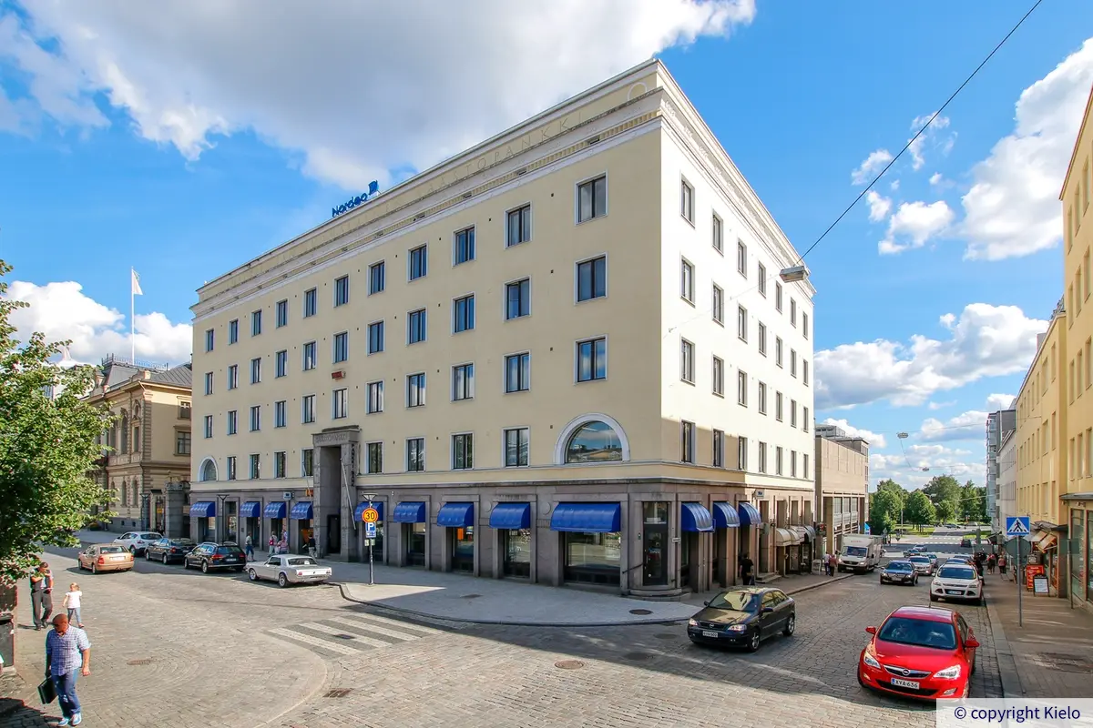 Hämeenlinnan Sibeliuksenkatu 2, ulkokuva. Kiinteistöllä on vaaleankeltainen julkisivu, katutason ikkunoissa on siniset markiisit. Rakennuksen ympärillä autoja pysäköitynä.