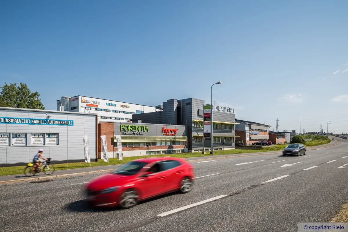 Oulun Voudintie 4, ulkokuva. Kiinteistön edustalla kulkee vilkas tie, rakennuksen seinällä on mainosbanderolleja ja yrityksien nimikylttejä.