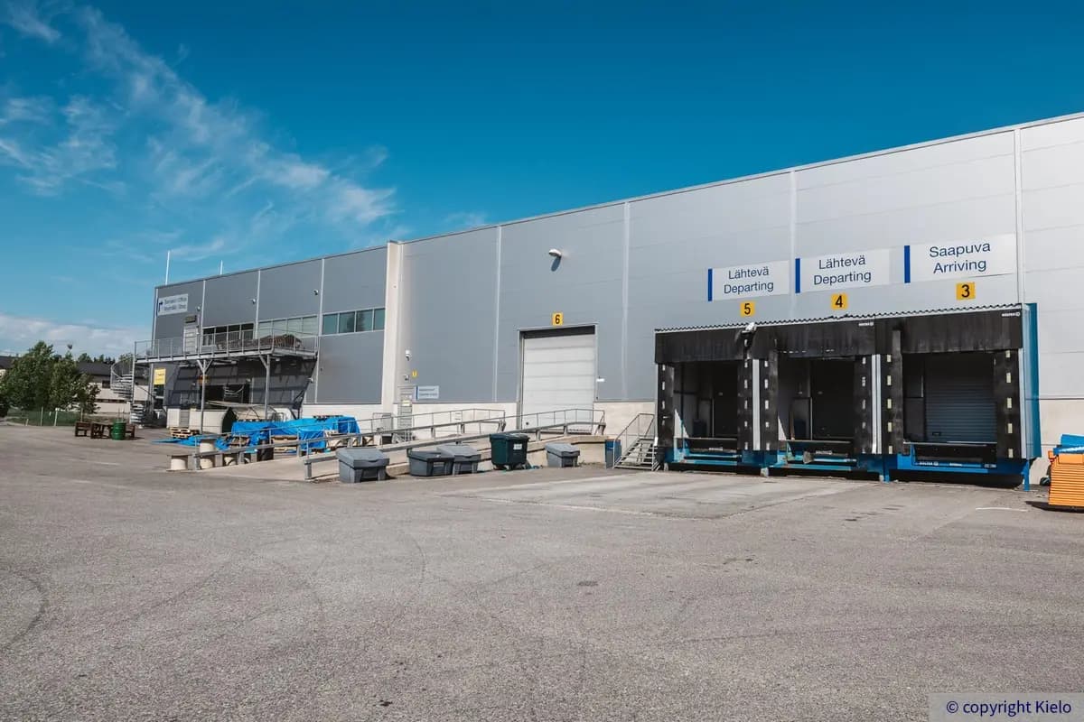 Kaarinan Asessorinkatu 3, ulkokuva. Kuvassa kolme lastaustaskua ja yksi nosto-ovi. Kiinteistön julki-sivu on harmaa, piha on asfaltoitu.