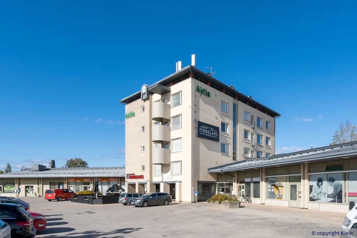 Tuusulan Hyrylänkatu 8, ulkokuva. Vaalea rakennus jonka kyljettä yrityksen nimikylttejä. Rakennuksen molemmilla puolilla matalammat osat joissa liiketiloja, edustalla pysäköintialue jossa autoja.