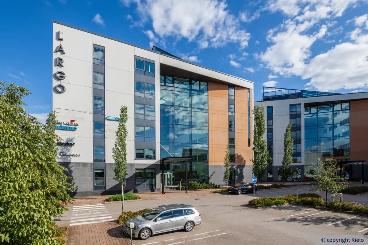 Gate8 Business Park Largo-toimitilakiinteistön julkisivu. Uudehkossa rakennuksessa on korkea lasiseinä sisäänkäynnin yläpuolella. Etualalla on pysäköintialue.