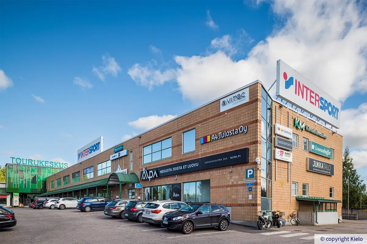 Tourukeskus ruskea kaakeloitu julkisivu. Seinustoilla runsaasti valomainoksia. Ensimmäisen kerroksen ikkunoiden yllä vihreät katokset.