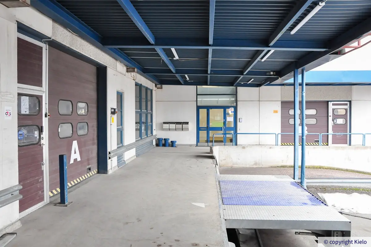 Helsingin Lirokuja 4 lastauslaituri sinisen katoksen alla. Lastauslaiturilta on nosto-ovet ensimmäisen kerroksen tiloihin, kulmasta ovi rappukäytävään.