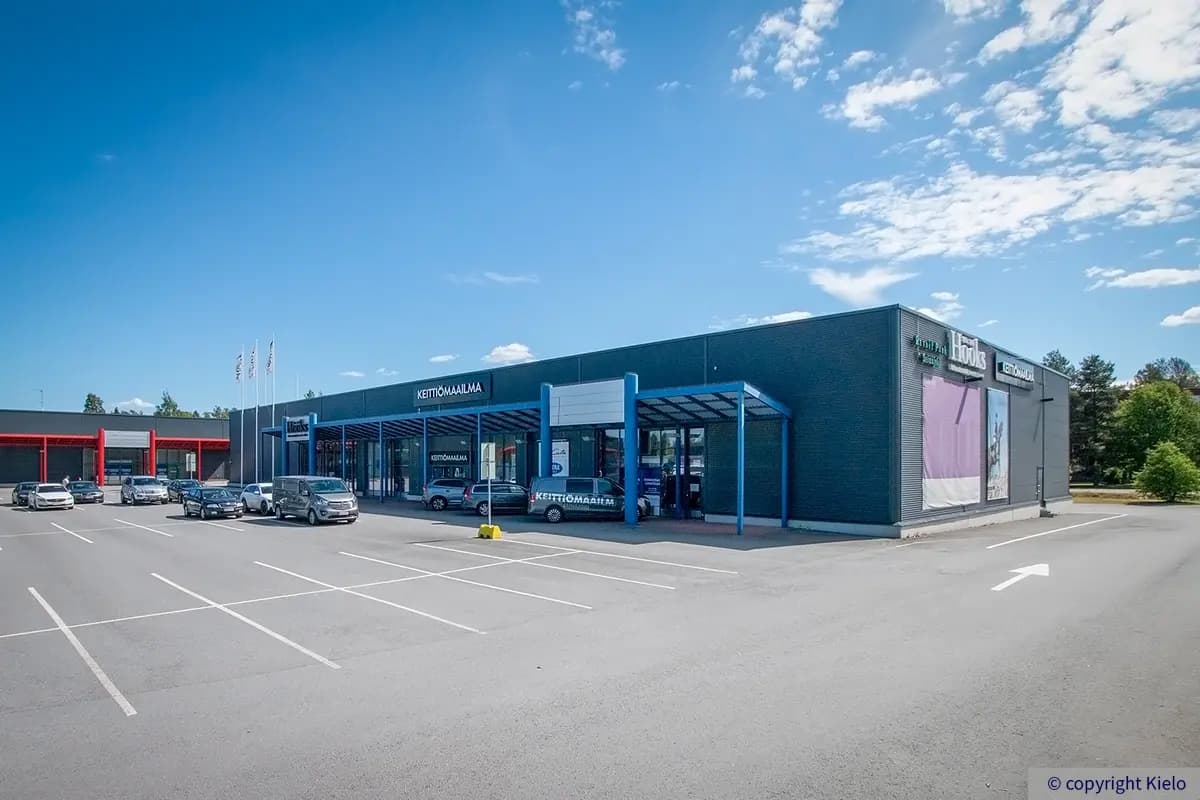 Seinäjoen Päivölänkatu 2, pienempi liikerakennuksista. Talon edessä on katettu, sinisin pylväin varustettu alue, jolta on kulku liiketiloihin.