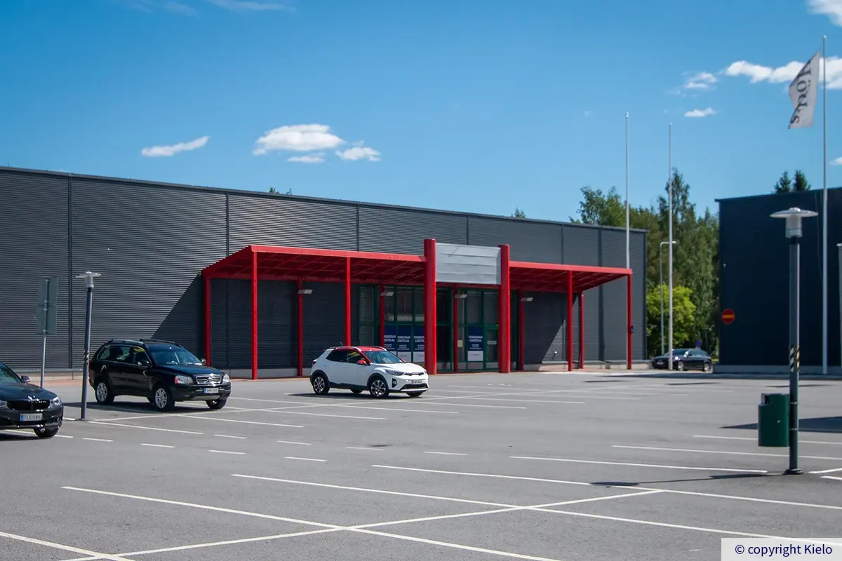 Seinäjoen Päivölänkatu 2, harman liikarakennuksen eteläpääty. Liiketilan edessä on punaisin rakentein katettu sisäänkäyntialue.