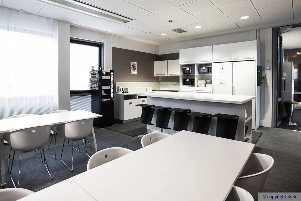 Jyväskylän Mattilanniemi 6-8 sijaitsevan 1. kerroksen 1260 m2 kokoisen toimitilan keittiötila, jossa valkoiset ruokapöydät. Keittiössä valkoiset kalusteet ja saareke, jonka ympäillä baarijakkaroita.