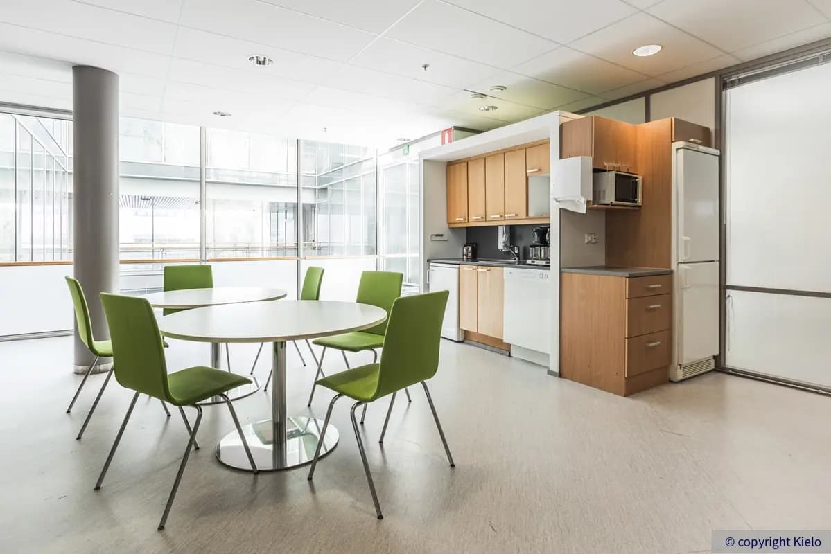 Jyväskylän Mattilanniemi 6-8 sijaitsevan toimitilan keittiö, jossa ruskeat kaapistot. Keittiön edessä kaksi pyöreää ruokapöytää, joiden ympärillä vihreitä tuoleja.