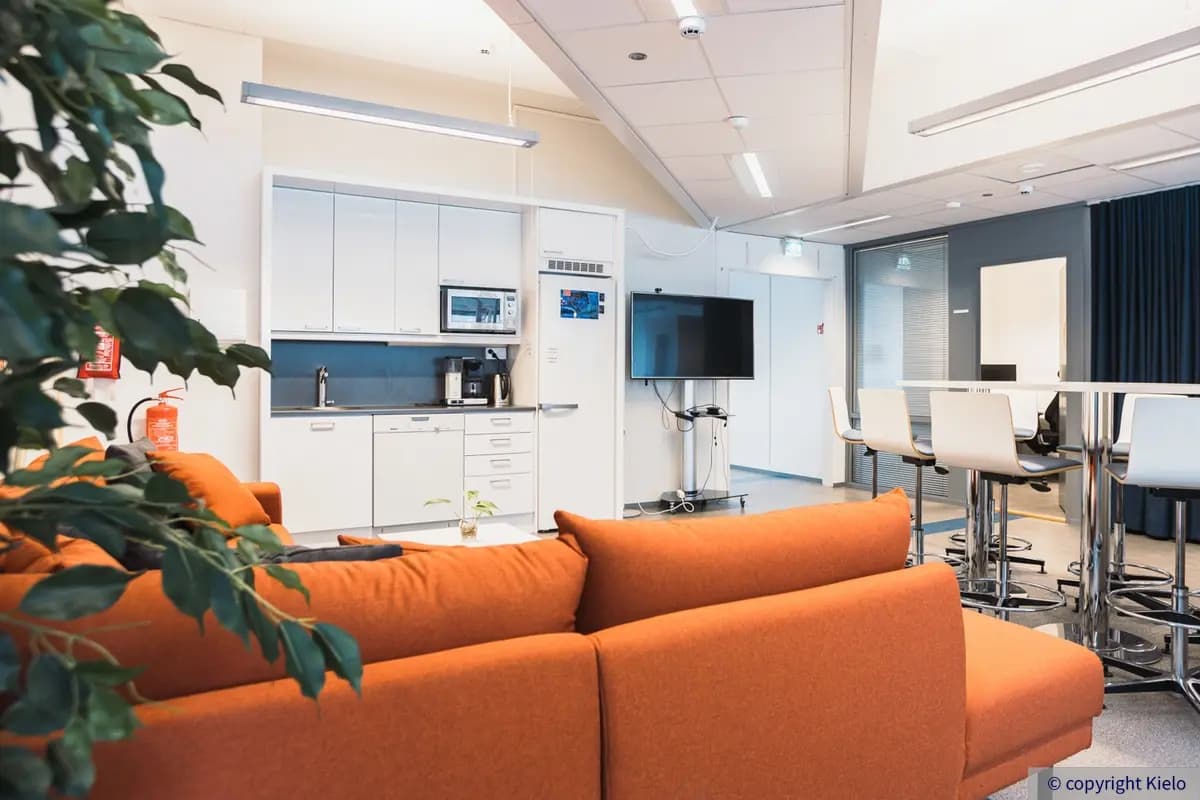 Innova 2 kiinteistön 3. kerroksen toimitilan keittiö, jossa iso oranssi kulmasohva ja valkoinen pyöreä ruokapöytä. Keittiössä jääkaappipakastin, astianpesukone ja mikro.