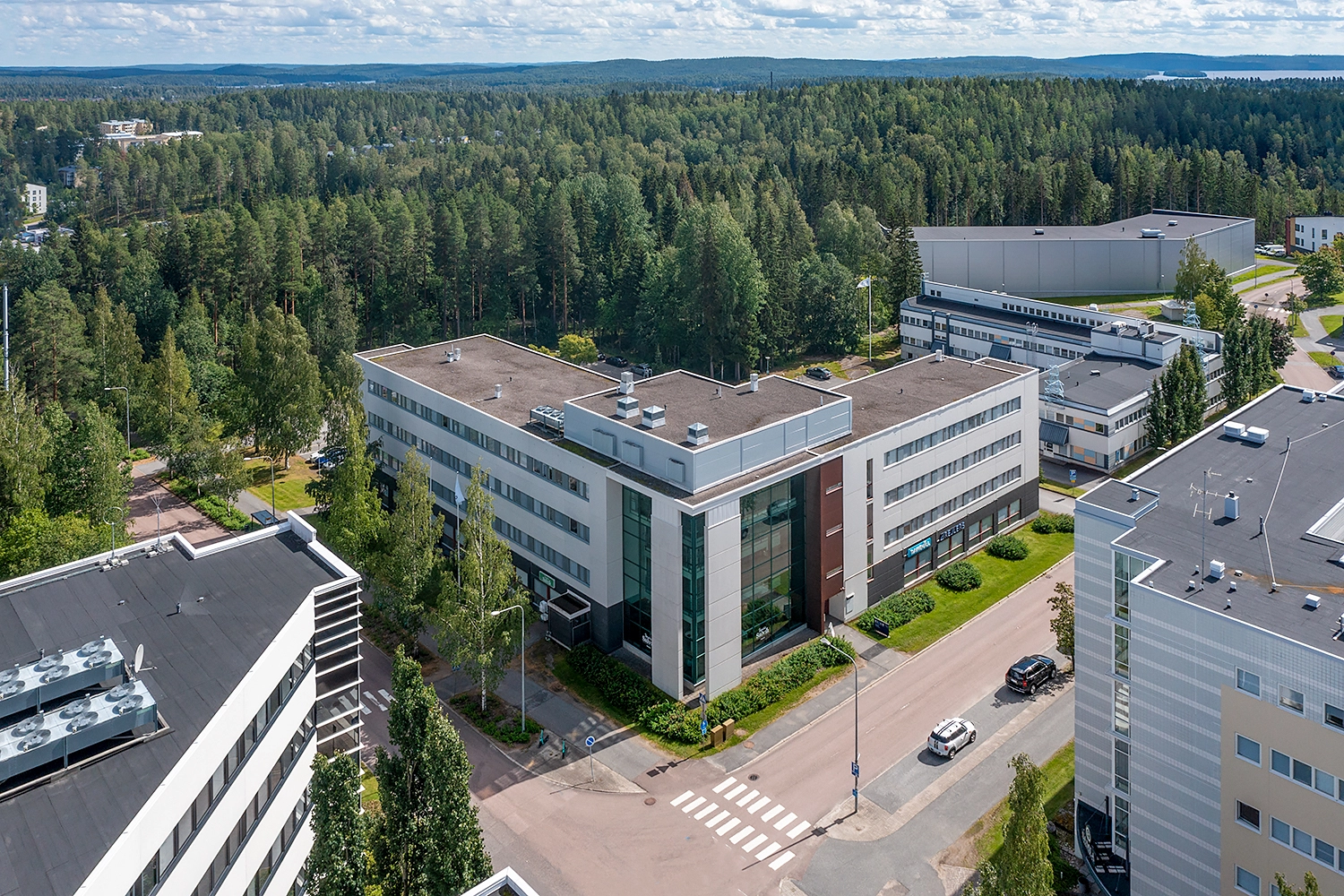 Toimitilakiinteistö Jyväskylän Ylistönmäentie 26 kuvattuna ilmasta. L-muotoinen 4-kerroksinen toimistotalo, jossa on tasakatto ja kulmassa lasinen atrium. Taustalla metsää. 