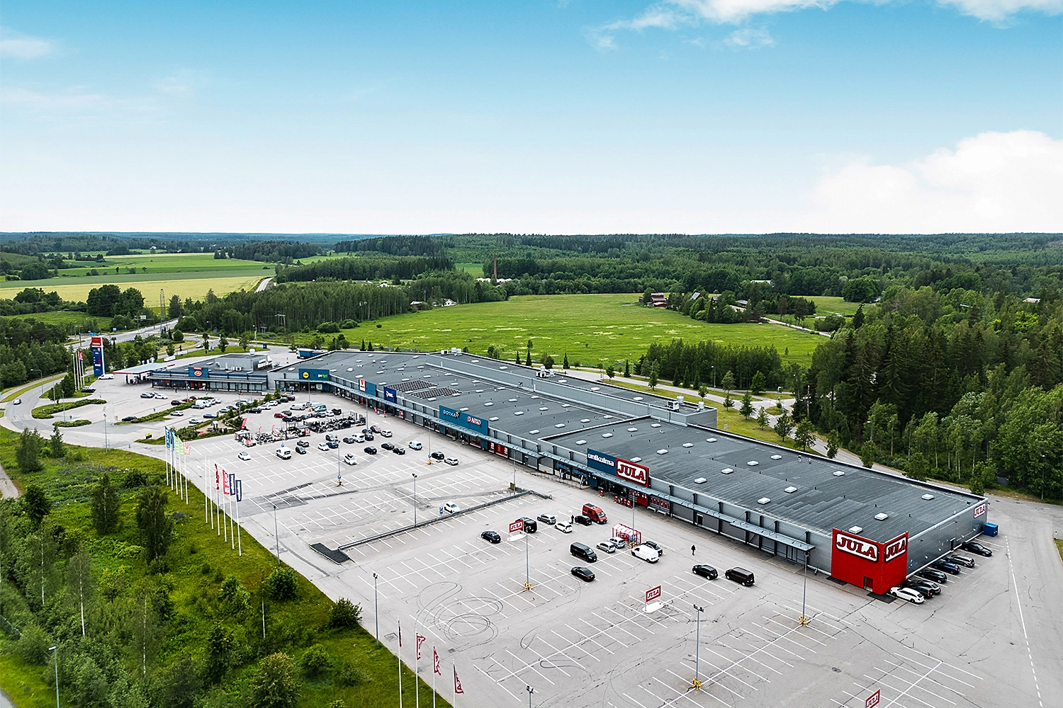 Kodinkeskus Sveitsin Portaali liikekiinteistö, jonka edessä suuri pysäköintialue. Rakennuksen takana metsä- ja peltomaisemaa.