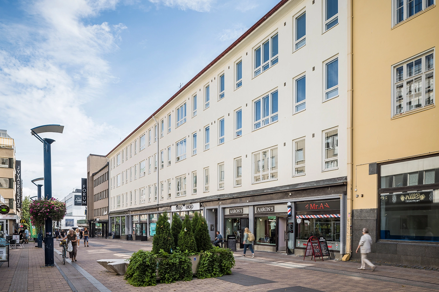 Jyväskylän Kauppakatu 18 vaalea julkisivu. Nelikerroksisen rakennuksen katutasossa on liiketiloja kävelykadun varrella.